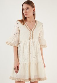 Robe crème avec un décolleté en V, corsage brodé, manches bouffantes et ourlets à franges. Présente des découpes décoratives et un tissu texturé.