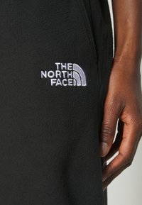 Svart fleece-tyg med mjuk textur och rektangulära fickor, med en vit broderad logotyp av "The North Face" på sidan.