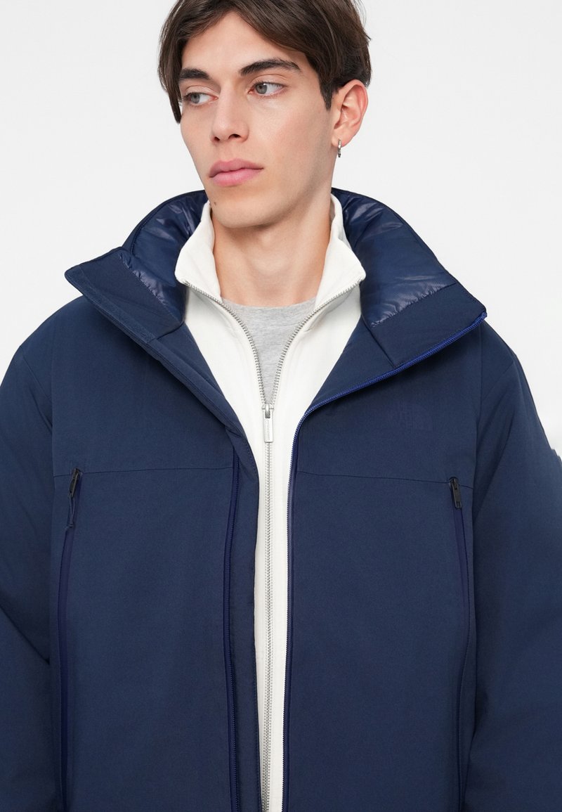 Doudoune bleu marine avec un col haut, comportant des poches zippées et une couche intérieure blanche zippée. Texture extérieure lisse, marque minimale.