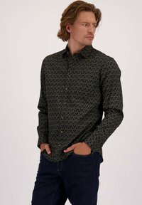 Man met lichtbruin haar, gekleed in een donker button-up shirt met een groen wijnklim patroon en een donkere spijkerbroek, met handen in zijn zakken, kijkend opzij.