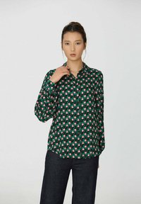 Blouse à manches longues et à boutons, de couleur noire, verte et rose avec un motif géométrique ; elle possède un col et une coupe décontractée, fabriquée en tissu léger.