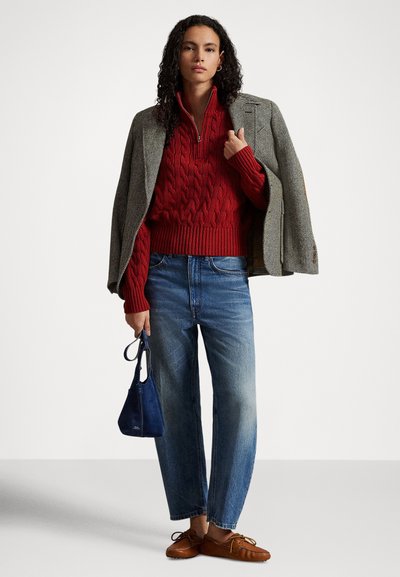 Γυναίκα που στέκεται φορώντας γκρι σακάκι πάνω από κόκκινο πλεκτό πουλόβερ με φερμουάρ, μπλε τζιν, καφέ μοκασίνια και κρατώντας μια μικρή μπλε τσάντα χειρός.