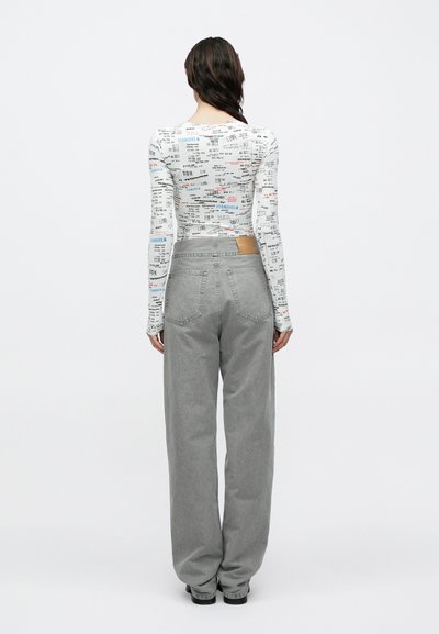 Dlhý rukáv biely top s viacfarebným textom a vzormi; v kombinácii s voľnými sivými nohavicami s koženou nášivkou na pásovke.