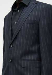 Homme portant une veste de costume bleu marine à fines rayures avec un revers cranté par-dessus une chemise noire à boutons, fermeture à deux boutons et poches à rabat.