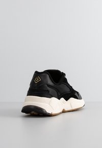 Baskets de sport noires et beiges avec une semelle épaisse et un logo au talon, photographiées de l'arrière sur fond gris.