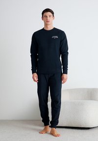 Černá dlouhá trička s malým logem na prsou, spojená s odpovídajícími černými kalhotami. Tkanina vypadá měkce a elasticky. Model stojí bos.