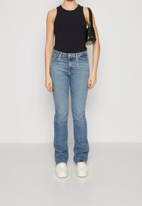 Donna che indossa un top senza maniche nero, jeans azzurri chiari, sneaker bianche e porta una borsa a tracolla nera su uno sfondo neutro.