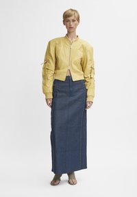 Geel bomberjack met geribbelde manchetten en ritszakken, gecombineerd met een lange blauwe denim rok met zijstuds. Het model draagt beige sandalen.