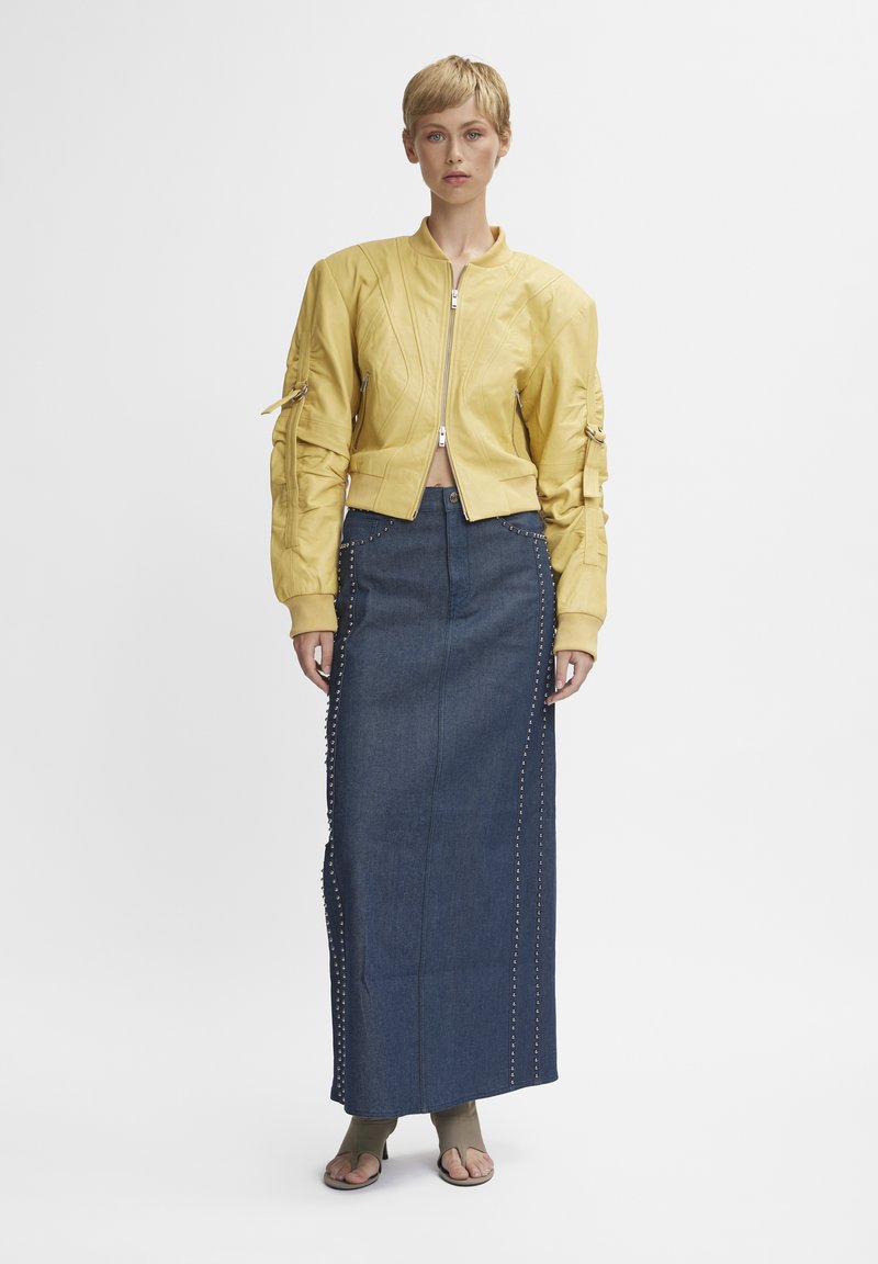 Veste bomber jaune avec des poignets côtelés et des poches zippées, associée à une longue jupe en denim bleu avec des clous sur les côtés. Le modèle porte des sandales beiges.