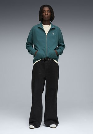 Persoon met dreadlocks tot op de schouders, een teal kleurige jas met rits, een wit shirt, zwarte wijd uitlopende broeken, witte schoenen en een ketting.