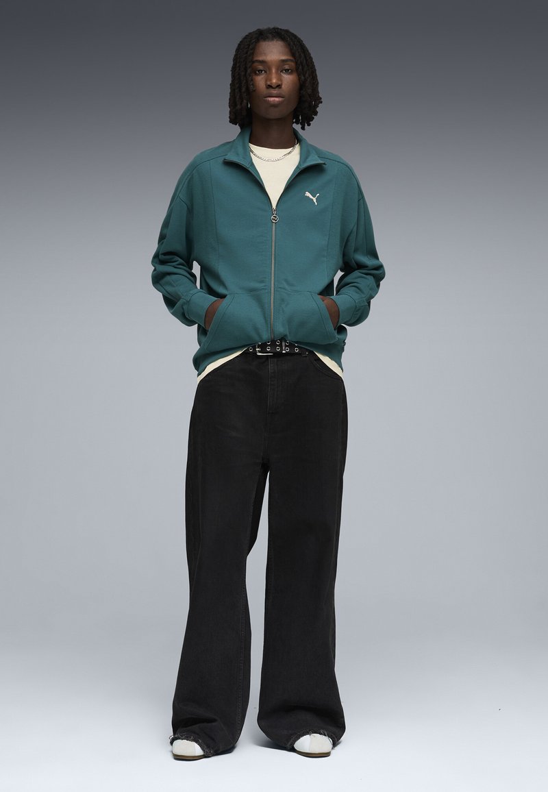 Personne aux dreadlocks mi-longues portant une veste zippée turquoise, une chemise blanche, un pantalon large noir, des chaussures blanches et un collier chaîne.