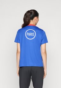 Mėlyna marškinė su raudonu apykakle, ant nugaros su apvaliu baltu logotipu su užrašu "PARIS". Pagaminti iš lygaus, lengvo audinio. Trumpomis rankovėmis.
