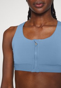 Sutiã desportivo azul com decote redondeado, detalhe de fecho na frente e faixa elástica na parte inferior. Tecido suave, design ajustado.