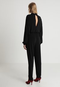 Svart jumpsuit med långa ärmar, öppen ryggdesign, elastisk midja och lös passform; matchad med röda högklackade skor.