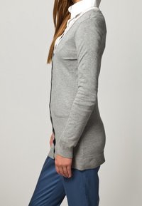 Grå cardigan med knäppning fram, ribbade ärmslut och fickor. Bärs över en vit skjorta med krage. Slät textur, figurnära design och knälång.