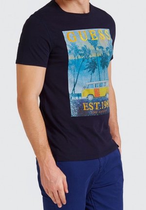 Man draagt een donkerblauw T-shirt met een kleurrijke strandscène, palmbomen, een gele bestelwagen en de tekst "GUESS EST. 1981 LOS ANGELES".