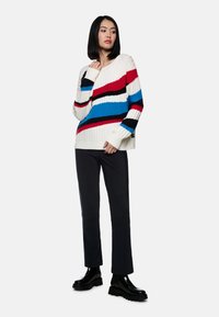 Strickpullover in Creme mit diagonalen Streifen in Rot, Blau und Schwarz; gerippte Textur. Kombiniert mit schwarzen Slim-Fit-Hosen und glänzenden schwarzen Stiefeln.