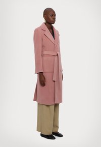 Lång rosa rock med bälte, gjord av mjukt material. Har en krage, två framfickor och rak skärning. Bärs med beige byxor.