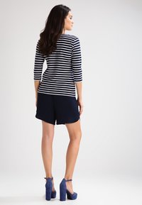 Marynarska bluzka w biało-niebieskie paski z rękawami 3/4, zestawiona z granatowymi szortami. Modelka ma na sobie niebieskie szpilki. Skupienie na swobodnym, luźnym kroju.