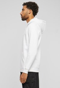 Sweatshirt blanc avec poche avant, capuche à cordon de serrage, poignets côtelés et texture lisse. Porté sur un pantalon foncé, vu de profil.