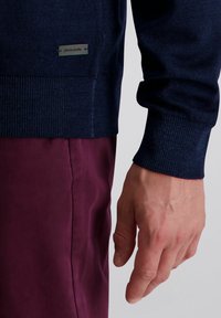 Dunkelblau gestrickter Pullover mit gerippten Bündchen und Saum. Mit einem silbernen Logo-Tag versehen. Kombiniert mit burgunderfarbenen Hosen, die eine glatte Textur zeigen.