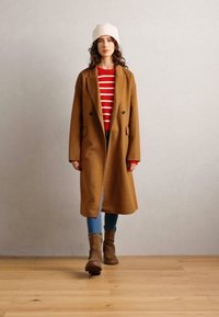 Brązowy wełniany płaszcz, sweter w czerwono-białe paski, niebieskie dżinsy i brązowe botki za kostkę. Biała czapka beanie, naturalnie falujące włosy, neutralne tło.