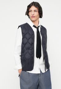 Ναυτικό υφασμάτινο γιλέκο με φερμουάρ μπροστά, με σχέδιο και όγκο. Φοράει πάνω από άσπρο πουκάμισο και μαύρη γραβάτα, με φαρδιά ανοιχτό μπλε παντελόνια.