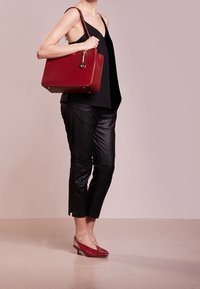 Punainen nahkainen käsilaukku, jossa on jämäkkä muotoilu, ketjuhihna ja kultaiset metalliosat. Malli pukeutunut mustaan toppiin ja lyhyisiin nahkahousuihin, jalassa punaiset korkokengät.