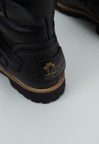Bota de cuero negro con un diseño robusto, que presenta un logo en relieve dorado en el talón, costuras blancas y una suela de goma gruesa.