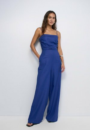 Femme debout contre un mur blanc portant une combinaison bleu sans manches à jambes larges, avec une main dans la poche et un bracelet argenté au poignet.
