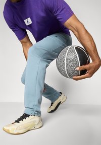 Lilla T-shirt med logo, lyseblå sweats, og beige sneakers, holder en sort og hvid mønstret basketball.