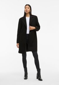 Cappotto di lana nero con revers, chiusura a un bottone e due tasche frontali, indossato su una camicia bianca e jeans scuri, abbinato a stivali neri.