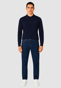 Polo a maniche lunghe blu navy con tre bottoni, logo sul petto; abbinato a jeans blu e sneakers bianche, creando un outfit casual.
