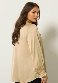 Beżowa bluzka z długimi, bufiastymi rękawami, miękką teksturą i zb gathers cuffs. Z okrągłym dekoltem i luźnym krojem.