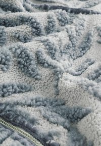 Tejido de forro polar suave y texturizado con un patrón de hojas en gris y blanco, mostrado en detalle de cerca con una cremallera parcialmente visible.