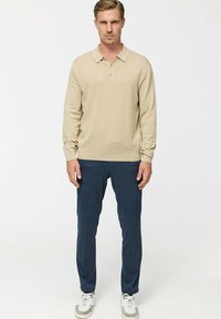 Un polo bej cu mâneci lungi dintr-un material tricotat moale, cu guler și placket cu nasturi. Asortat cu o pereche de pantaloni închiși la culoare, concepuți pe linie conică, și adidași albi.