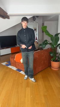 Young man in dark jacket and jeans stands in living room with orange couch, large green plant, wooden floor, and television.