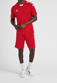 Červený sportovní dres a šortky vyrobené z prodyšné látky; obsahují bílé akcenty na ramenou a logu; v kombinaci s bílými teniskami.