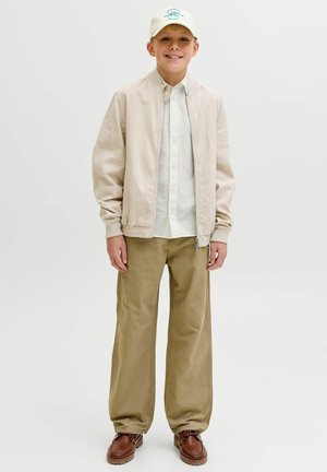 Jongen draagt een beige bomberjack, wit shirt, kaki broek, bruine schoenen en een witte pet, staat en glimlacht tegen een effen lichte achtergrond.