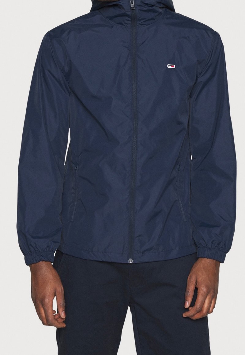 Homme portant une veste à capuche bleu marine à fermeture éclair avec des poignets élastiques et un petit logo sur la poitrine gauche, mains reposant le long du corps.
