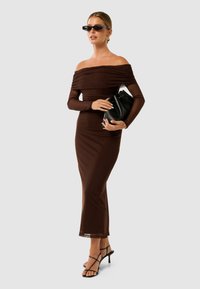 Robe marron dénudée des épaules en tissu extensible, cintrée à la taille, avec un design épuré. Associée à des talons noirs à lanières et à une pochette noire.