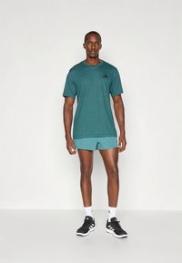 Turkos fukttransporterande sport-T-shirt med korta ärmar och en diskret svart logotyp på bröstet, kombinerad med matchande turkosa shorts och svarta sneakers.