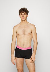 Fekete pamut boxer alsónadrág pink Lacoste logós derékpánttal. A modellen nincs póló, közepes szőrtelítettségű testtel és semleges arckifejezéssel látható.