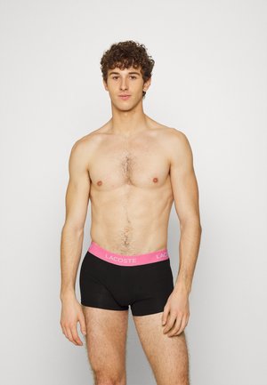 Schwarze Baumwoll-Boxershorts mit einem pinken Lacoste-Logo-Bund. Das Model ist Oberkörperfrei und zeigt feine Körperhaare sowie einen neutralen Gesichtsausdruck.