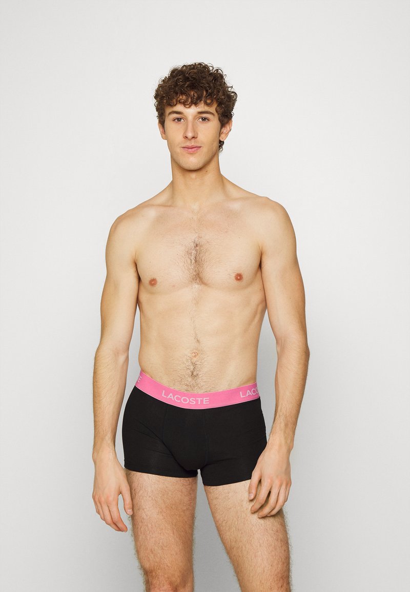 Fekete pamut boxer alsónadrág pink Lacoste logós derékpánttal. A modellen nincs póló, közepes szőrtelítettségű testtel és semleges arckifejezéssel látható.