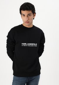 Crna dukserica od mekog materijala, s rebrastim manžetama i izrezom, te bijelim otisnutim logotipom: "KARL LAGERFELD 21 RUE ST-GUILLAUME PARIS."