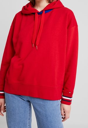 Personne portant un sweat à capuche rouge vif avec cordons et un jean bleu clair, vue des épaules aux hanches, debout devant un fond uni.