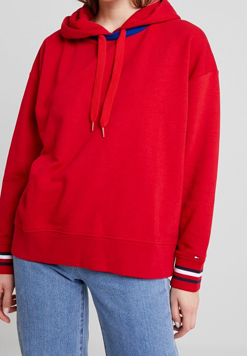 Personne portant un sweat à capuche rouge vif avec cordons et un jean bleu clair, vue des épaules aux hanches, debout devant un fond uni.