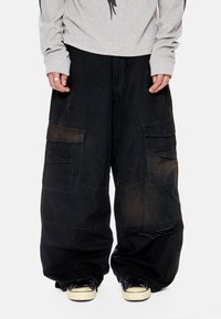 Άτομο που φοράει υπερμεγέθη μαύρο παντελόνι cargo με μεγάλες τσέπες και σκίσιμο στο γόνατο, συνδυασμένο με μαύρα αθλητικά παπούτσια και γκρι ανοιχτό μακρυμάνικη μπλούζα.