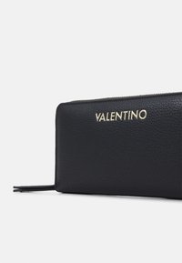 Juodas odinis piniginė su tekstūruota paviršiumi, auksiniu "Valentino" logotipu ir užtrauktuku viršutinėje kraštinėje.